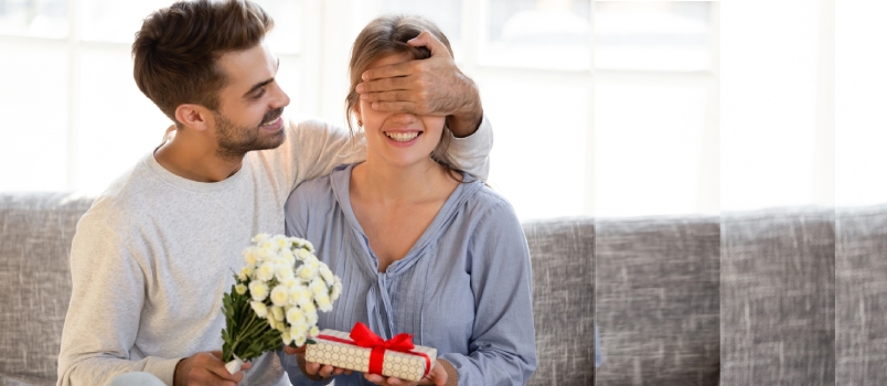 Hombre amoroso esposo novio con flores cerrando los ojos de mujer novia sosteniendo caja de regalo adivinando presente haciendo sorpresa romántica a esposa