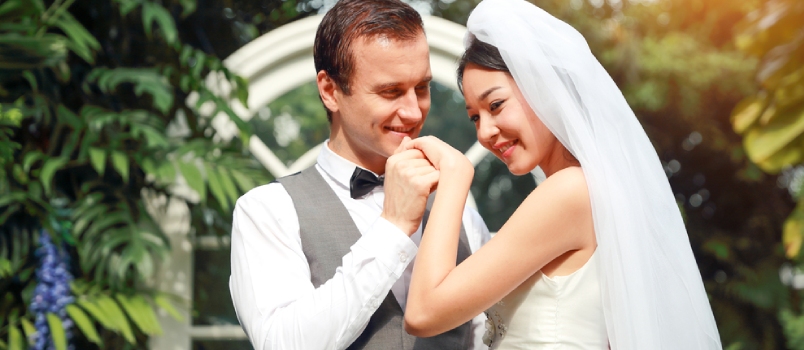 El guapo novio caucásico sostiene y besa la mano de la novia asiática con amor y cara sonriente durante la boda