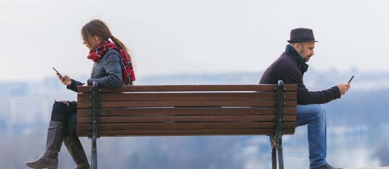 Hombre y mujer sentados aparte en un banco del parque y usando el móvil