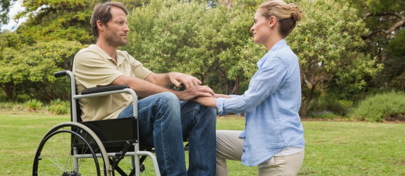 Hombre contento en silla de ruedas con su pareja arrodillada a su lado y mirándose en un parque